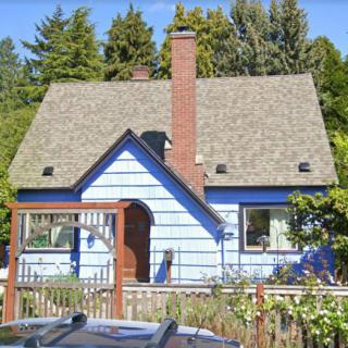 A charming blue cottage with a chimney and garden, surrounded by trees.