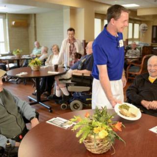 A staff member serves food to residents in a nursing home dining area.