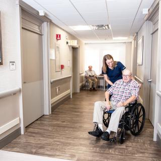 A caregiver assists an elderly man in a wheelchair down a hallway in a nursing home.