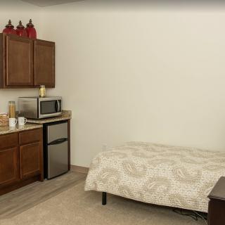 A small apartment room featuring a bed and a kitchenette with cabinets, a microwave, and a mini fridge.