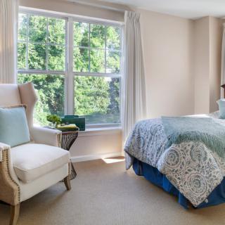 A cozy bedroom with natural light, featuring a plush armchair and a neatly made bed near a large window.
