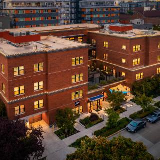 Aerial view of a brick building at dusk with lights on, surrounded by trees and parked cars.