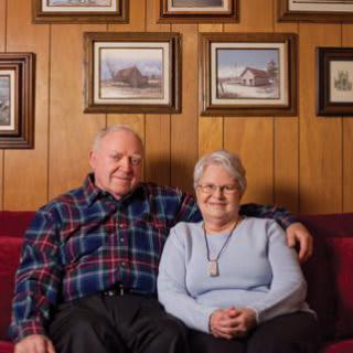 An elderly couple sitting together on a couch in their living room, surrounded by framed pictures.