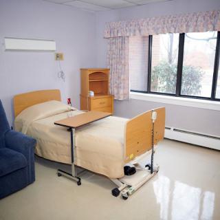 A clean and simple hospital room with a single bed, chair, and window providing natural light.