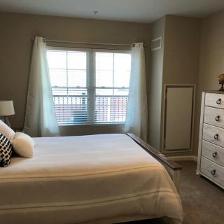 A cozy bedroom with a neatly made bed, a dresser, and a large window letting in natural light.
