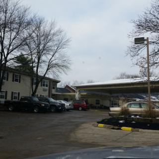 A view of a parking area with several cars parked near two buildings under a cloudy sky.