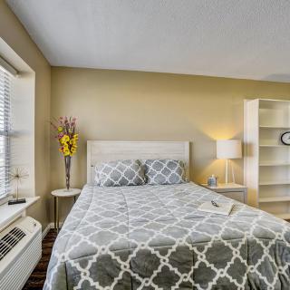 A cozy bedroom featuring a bed with patterned bedding, a lamp on a bedside table, a window providing natural light, and shelves for storage.