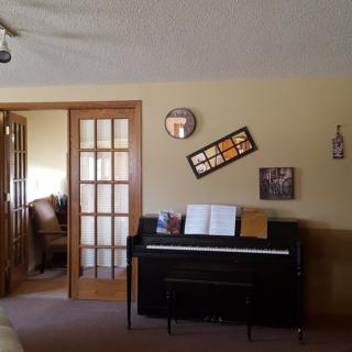 A cozy living room featuring a piano with sheet music, framed photos on the wall, and an open wooden door leading to another room.