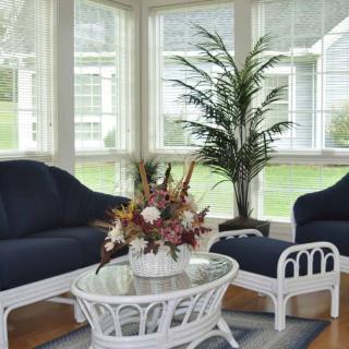 A bright sunroom with wicker furniture and a potted plant, featuring large windows and a glass coffee table.