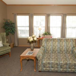 A cozy living room featuring a patterned sofa, armchair, and coffee table with decorative plants and ample natural light from the windows.