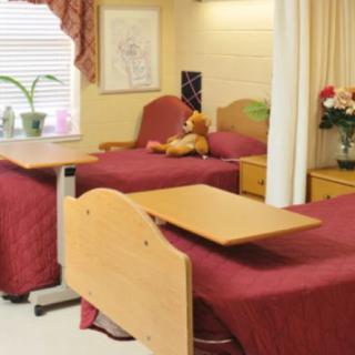 A hospital room with two beds, each covered in red bedspreads, featuring overbed tables, a teddy bear, and decorative elements like flowers and curtains.