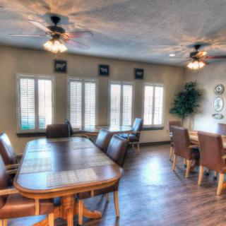 A spacious dining room with wooden tables and chairs, illuminated by ceiling lights and natural light from large windows.