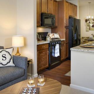 A cozy apartment interior featuring a living room with a sofa and lamp, adjacent to an open kitchen with wooden cabinets.