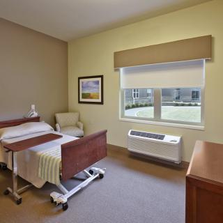 A simple and clean hospital room with a bed, bedside table, chair, and a window overlooking a garden.