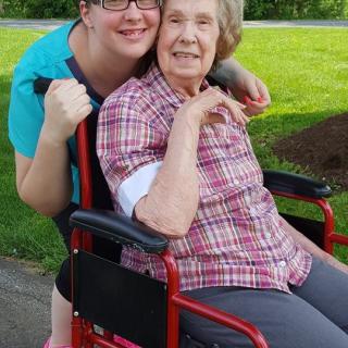 A younger woman hugging an elderly woman in a wheelchair outdoors.