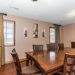 A dining room with a large wooden table surrounded by chairs, featuring windows with curtains and decorative wall art.