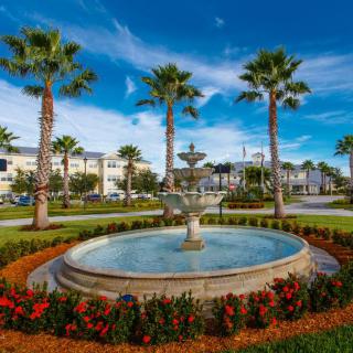 A beautifully landscaped area featuring a central fountain surrounded by palm trees, with modern buildings in the background under a clear blue sky.