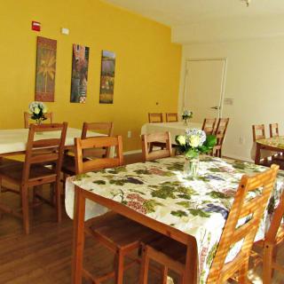 A cozy dining area featuring wooden tables and chairs, adorned with floral tablecloths against a yellow accent wall.