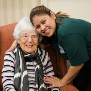 An elderly woman sitting and smiling with a caregiver beside her.