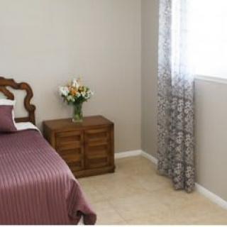 A cozy bedroom featuring a bed with a purple cover, a wooden nightstand with flowers, and patterned curtains by the window.