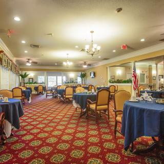 A spacious dining room with elegantly set tables covered in blue tablecloths and cushioned chairs, all resting on a patterned carpet under warm lighting from chandeliers.