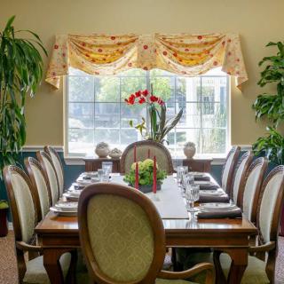 A well-decorated dining room with a large table and chairs, surrounded by plants and illuminated by a window.