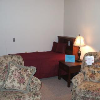 A cozy bedroom setup featuring a bed with burgundy bedding, a wooden nightstand with a lamp, and floral-patterned armchairs.