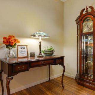 A cozy corner featuring an antique desk with a table lamp, vibrant flowers, and a classic grandfather clock.