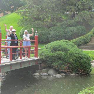 A group of people waving while standing on a bridge in a lush garden.