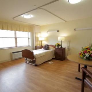 A clean and well-lit hospital room with a single bed, wooden flooring, and floral decorations.