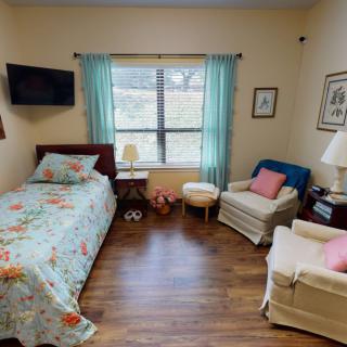 A cozy bedroom featuring a single bed, two armchairs, and a TV, with natural light streaming through the window.