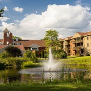 A serene landscape view featuring a fountain in a pond surrounded by lush greenery and buildings under a partly cloudy sky.