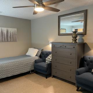 A cozy bedroom featuring a single bed, gray dresser, comfortable armchairs, and stylish decor.