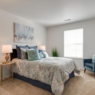 A cozy and elegantly decorated bedroom featuring a neatly made bed with patterned bedding, side tables with lamps, a piece of artwork above the bed, and a window letting in natural light.