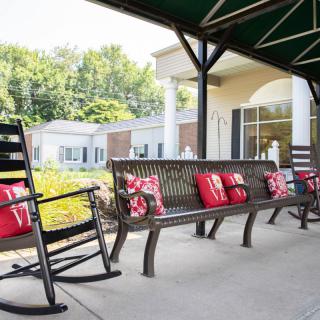 A cozy outdoor seating area featuring benches and a rocking chair adorned with red pillows, situated under an awning in front of a building.