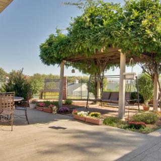 A sunny patio with a vine-covered pergola and outdoor seating, surrounded by well-maintained landscaping.