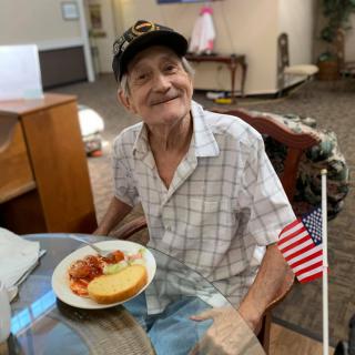 An elderly man wearing a hat smiles while sitting at a table with a meal and an American flag.