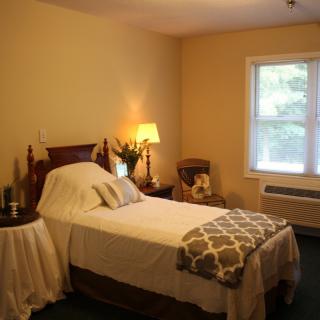 A cozy bedroom featuring a neatly made single bed, with warm lighting and a window overlooking greenery.