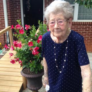 An elderly woman standing next to a pot of vibrant flowers in an outdoor garden setting.