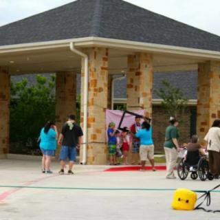 A group of people gathered outside a building, including someone in a wheelchair and a person carrying a cross.