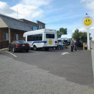 A small bus and several cars are parked in a parking lot near a building, with a smiley face sign visible against the blue sky.