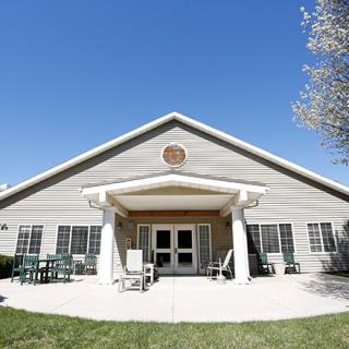 A single-story building with a gabled roof and a welcoming entrance surrounded by outdoor seating, set against a clear blue sky.