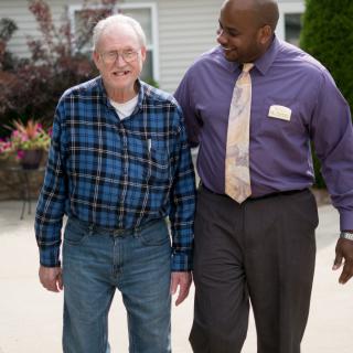 A caregiver assists an elderly man while walking outdoors.