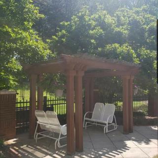 A wooden pergola providing shade over white outdoor chairs on a sunny patio.