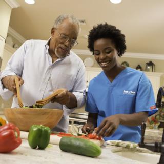 An elderly man and a caregiver are smiling while preparing vegetables together in the kitchen.