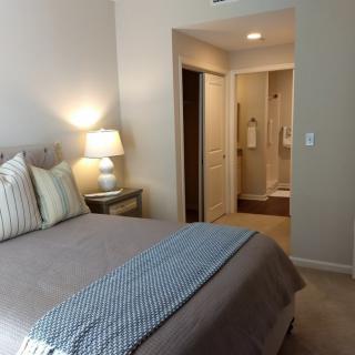 A cozy bedroom with neutral tones, featuring a neatly made bed, side lamp, and an open doorway leading to the bathroom.