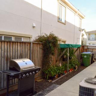 A backyard with a grill, a small garden area, and trash bins along a wooden fence.