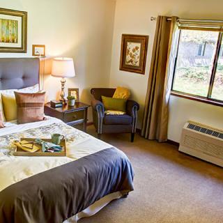 A cozy bedroom with a neatly made bed, side table, and a comfortable chair by the window.
