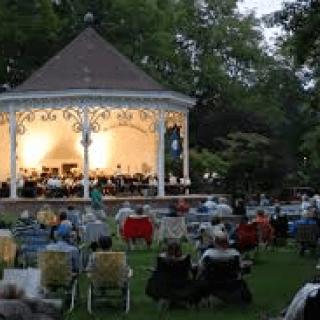 An outdoor evening concert in a park with an audience seated near a gazebo.