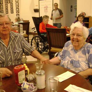 An elderly couple is sitting together at a dining table, holding hands in a cozy restaurant setting.